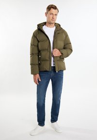 Oliven grøn pufferjakke med hætte, trykknapper og frontlommer. Båret over en hvid t-shirt, parret med blå jeans og hvide sneakers.