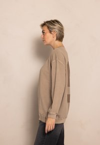 Beige sweatshirt with ribbed cuffs, round neckline, and subtle color blocking in soft brown tones. Worn over dark gray jeans.