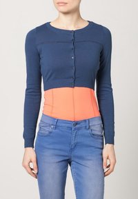 Marinblå kort kcardigan med knappstängning och ribbade manschetter över en orange åtsittande topp; kombinerat med ljusblå denimjeans.