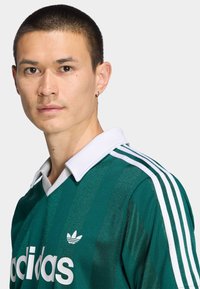 Πράσινη φανέλα με λευκές ρίγες, λευκό γιακά και ανάγλυφη υφή. Διαθέτει λευκό λογότυπο Adidas στο στήθος. Κοντά μανίκια.