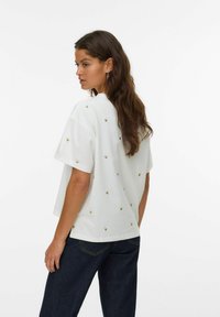 Hvid bomulds T-shirt med korte ærmer, prydet med gyldne hjertebroderier spredt over ryggen. Løs pasform, rund halsudskæring og slidser i siderne.