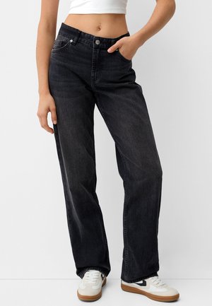 Frau trägt schwarze, hoch taillierte Jeans mit weitem Bein, kombiniert mit weißen Turnschuhen und einem weißen, bauchfreien Top, steht vor einem schlichten Hintergrund.