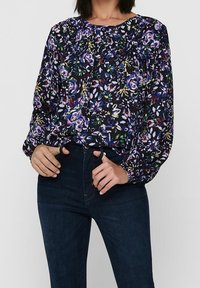 Blusa floreale con maniche lunghe, sfondo nero, motivi vivaci in viola e verde, dettagli arricciati sul colletto, abbinata a jeans scuri.