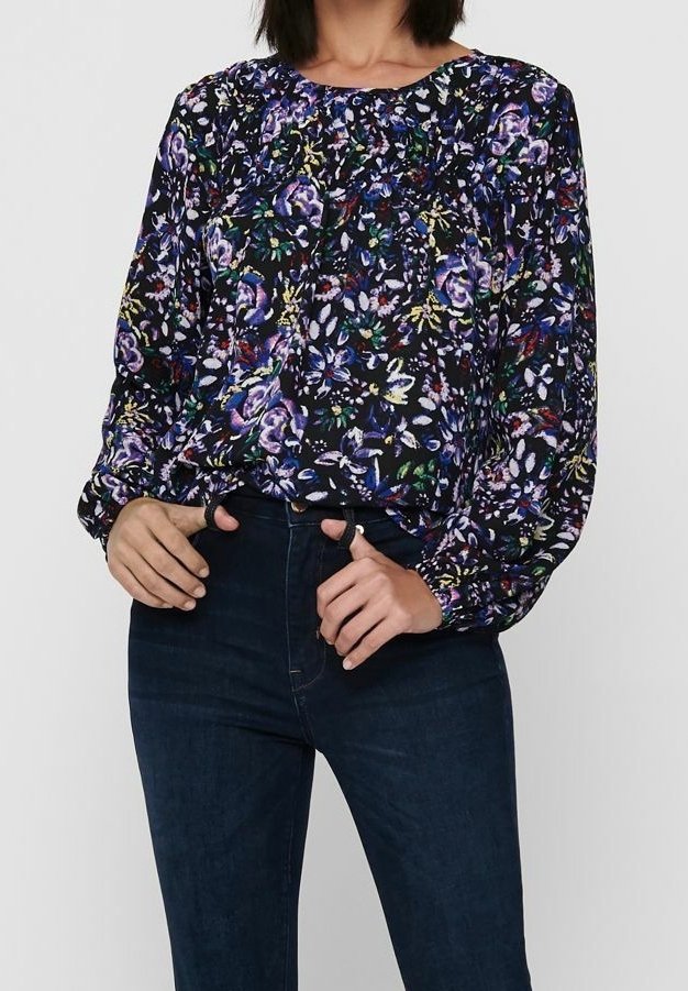 Blusa floreale con maniche lunghe, sfondo nero, motivi vivaci in viola e verde, dettagli arricciati sul colletto, abbinata a jeans scuri.