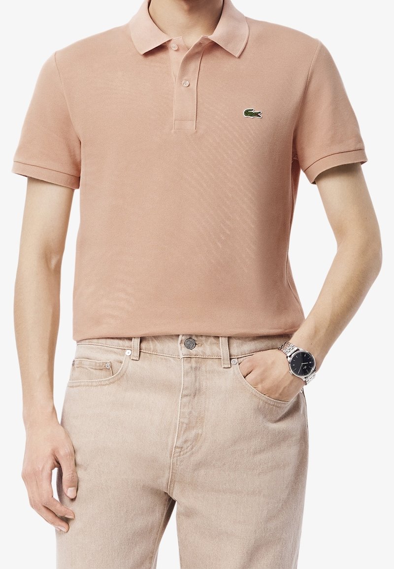 Homme portant un polo à manches courtes rose clair avec un logo de crocodile vert, un pantalon beige et une montre-bracelet argentée.