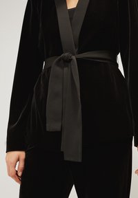 Czarna aksamitna marynarka z satynowym paskiem w talii. Posiada kołnierz szalowy i długie rękawy, prezentując miękką teksturę i elegancki design.