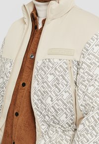 Puchowa kurtka w kremowym kolorze z wzorem po lewej stronie. Posiada naszywkę z logo oraz kieszeń na zamek, zestawiona z brązową koszulą z kordurowej tkaniny.