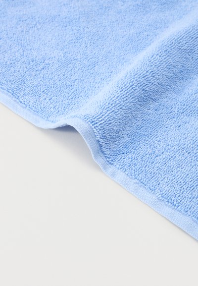 Modro svetel brisača iz frotirja z vidnimi zankastimi vlakni, obešena na belem površju, prikazuje šivano rob in rahlo gubo.