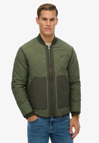 Gequilted bomberjack in donkergroen olijf met ribgebreide manchetten, twee voorzakken en een ritsluiting. Voorzien van een subtiel logo op de borst.