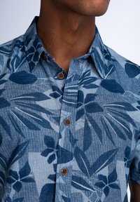 Camisa de botones con un estampado de hojas en azul y azul claro, con cuello, mangas cortas y botones de madera a lo largo de la tapeta.