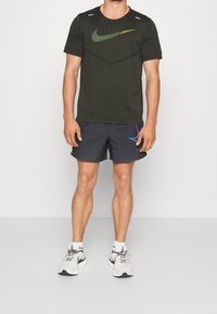 Camiseta de manga corta de Nike en verde oscuro con un logo en verde y naranja, combinada con pantalones cortos negros con un acento azul y zapatillas blancas.