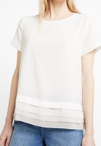 Top blanco de manga corta hecho de tela ligera, con un dobladillo en capas texturizado que presenta rayas horizontales en un tono ligeramente más oscuro.