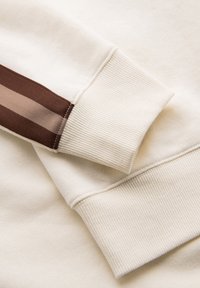 Beige sweatshirtærme med ribbet manchet og todelt brun stofstribe langs armen.