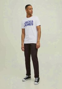White t-shirt with blue "JACK & JONES" text, paired with dark brown pants and black sneakers. Model standing against a light background.