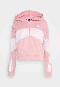 Sweat à capuche zippé rose et blanc Adidas avec poches avant, capuche réglable avec cordon et détails blancs à trois bandes sur les manches.