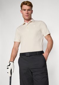 Beige Poloshirt mit kurzen Ärmeln und kleinem Logo, kombiniert mit schwarzen Hosen und einem schwarzen Gürtel. Weißer Golfhandschuh mit schwarzen Akzenten.