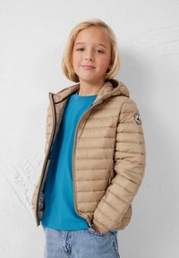 Lichtbeige gewatteerd jack met horizontale stiksels en een capuchon, gedragen over een turquoise shirt, met een logopatch op de linkermouw.