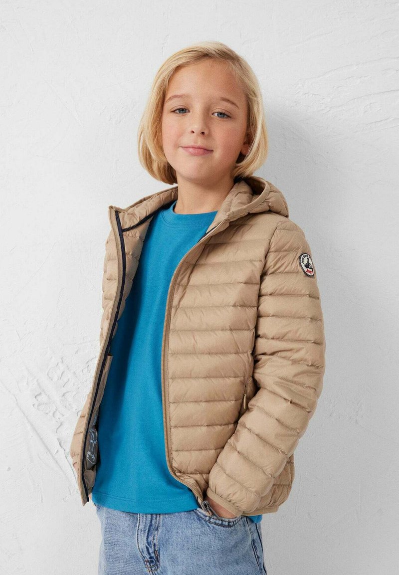 Lichtbeige gewatteerd jack met horizontale stiksels en een capuchon, gedragen over een turquoise shirt, met een logopatch op de linkermouw.