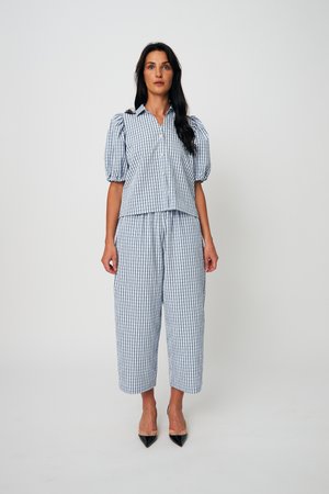 Femme debout portant une chemise à manches courtes boutonnée à carreaux bleu clair et blanc assortie à un pantalon large raccourci, avec des talons noirs pointus.