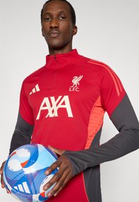 Rød Liverpool FC træningstrøje med halv lynlås, kontrasterende grå ærmer, hvidt logo og AAX-branding, parret med en blå og hvid mønstret fodbold.