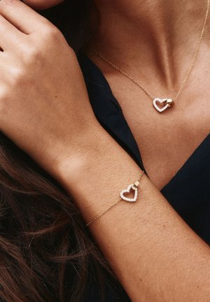 Kvinna bär guldfärgat halsband i form av ett hjärta och matchande armband med små diamanter, klädd i mörk topp, med handen vilande nära ansiktet.