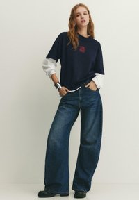 Junge Frau mit langen Haaren, die ein schwarzes "THE MET" T-Shirt über einem weißen Langarmshirt, weite Jeans, schwarze Schuhe und ein silbernes Armband trägt.