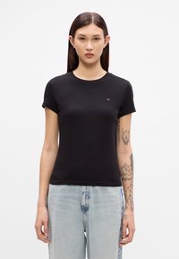 Svart bomull T-skjorte med korte ermer og rund hals. Har en liten, brodert logo på brystet. Kombinert med lyseblå jeans.