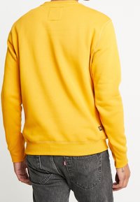 Sudadera amarilla con puños y dobladillo acanalados, hecha de una tela suave. Presenta un parche cuadrado en la parte posterior, cerca del escote.