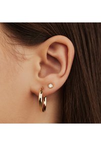 Goldene Creolen und ein kleiner Stecker mit einem klaren Stein, die eine glatte Textur und reflektierende Oberfläche gegen braunes Haar zeigen.