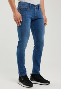 Mees, kes kannab kitsaid siniseid teksapükse ja musti tossusid, seisab ühevärvilise hele tausta ees, käed lõdvalt külgedel.