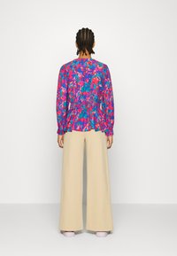 Blouse à manches longues dans un motif floral rose vif et bleu avec taille froncée. Associée à un pantalon large crème et des chaussures rose pâle.