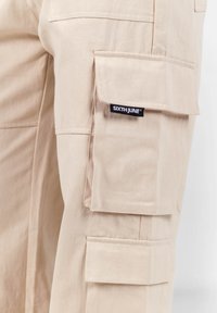 Beige cargobroek met meerdere grote zakken, gestikte details en een klein zwart label met "SIXTH JUNE" aan de zijkant.