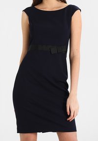 Mörkblå figurnära klänning med rundad halsringning, korta ärmar och ett svart band med en rosett i midjan. Slät tygtextur.