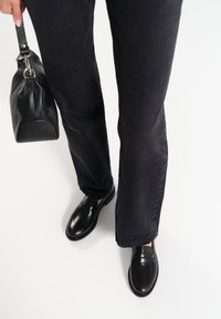 Zwarte denim jeans met een rechte pijp, gecombineerd met zwarte leren instappers. Een kleine zwarte leren handtas wordt aan de zijkant vastgehouden.