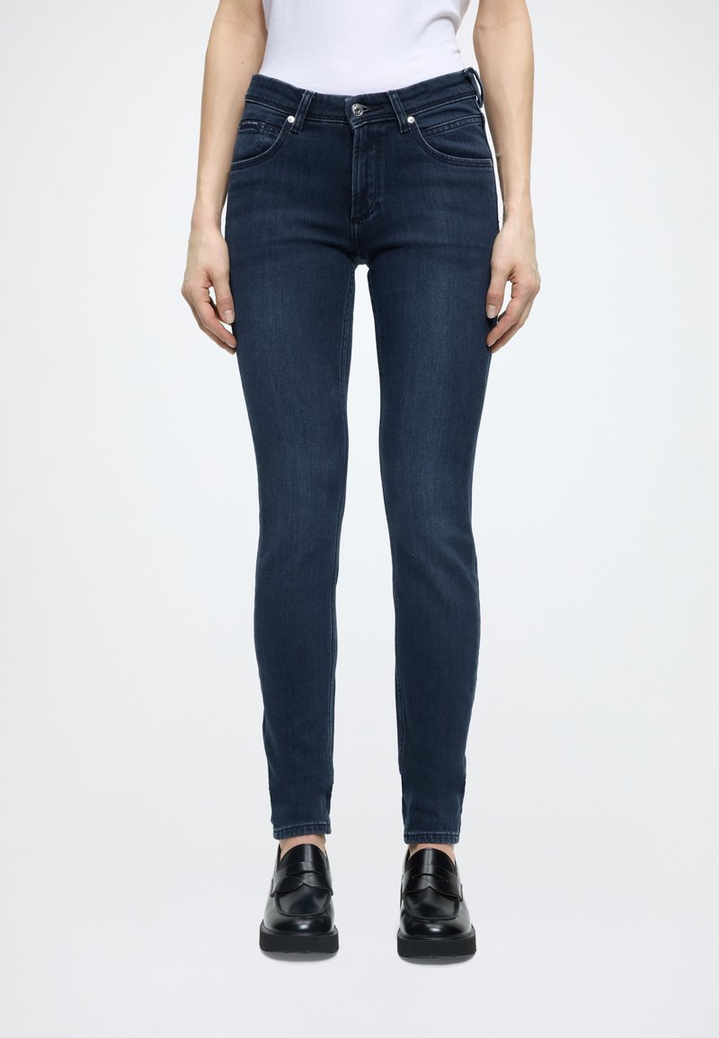 Tumši zilas šauras džinsa bikses ar augstu vidukli, piecām kabatām un sudraba aksesuāriem. Pasvītrotas ar melniem loaferiem.