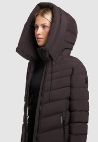 Vrouw in een donker gewatteerd jack met een oversized capuchon, rits dicht, kijkt opzij tegen een effen lichte achtergrond.