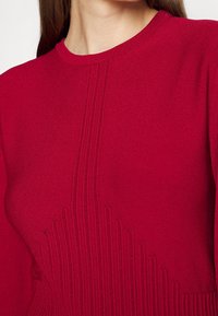 Roter Strickpullover mit geripptem Muster und vertikalen Streifen sowie einer figurbetonten Silhouette. Runder Halsausschnitt und lange Ärmel sind sichtbar.
