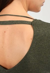 Khaki zelený top s hlubokým designem V-zády a dvěma tenkými ramínky. Měkká textura s jemným lesklým efektem a mírným skvrnitým vzorem.