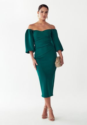 Kobieta ubrana w turkusową sukienkę midi z odkrytymi ramionami i bufkami na rękawach, trzyma plecioną złotą kopertówkę i nosi złote szpilki z paskiem wokół kostki.