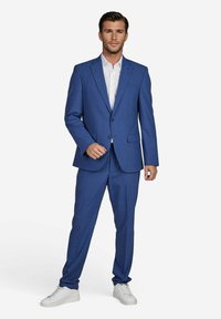 Blaues maßgeschneidertes Anzug mit Reverskragen, Ein-Knopf-Verschluss und passenden Hosen. Getragen über einem weißen Hemd und kombiniert mit weißen Turnschuhen.