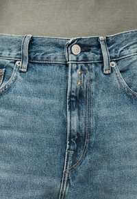 Κοντινή λήψη της μέσης μπλε τζιν με μεταλλικό κουμπί, θηλιές για ζώνη και μπροστινές τσέπες πάνω σε γκρι υφασμάτινο φόντο.