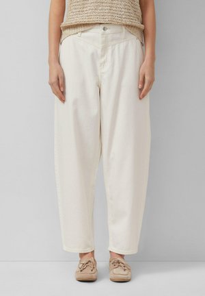 Kobieta ubrana w luźne, kremowobiałe spodnie, beżowy sweter i beżowe buty w stylu mokasynów, stojąca na tle jednolitego jasnoszarego tła.