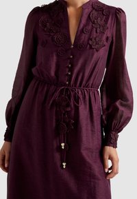Šaty s dlhým rukávom v tmavofialovej farbe s kvetinovou výšivkou, zapínaním na gombíky a obtiahnutým pásom. Majú textúrovanú látku a dekoratívne šnúrky na leme.
