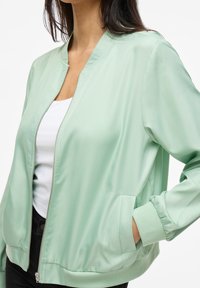 Giacca bomber verde menta realizzata in tessuto liscio e leggero. Presenta una chiusura con zip, polsini a costine e tasche laterali. Design dalla vestibilità standard.