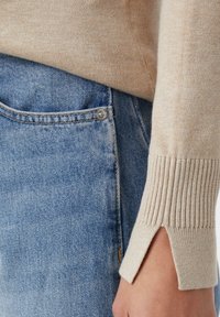 Beige gerippter Pulverschopf mit einem kleinen Schlitz, der über dem Taschenbereich von hellblauen Jeans getragen wird.