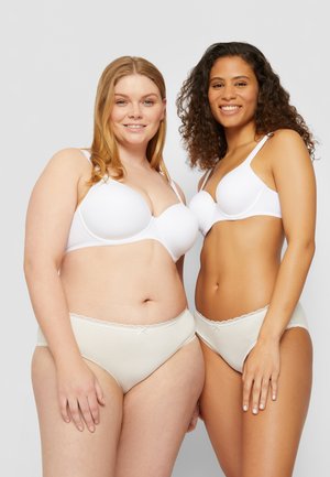 Dos mujeres sonrientes de pie una al lado de la otra, vistiendo sujetadores blancos y ropa interior beige, frente a un fondo gris liso.