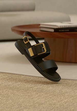 Sandale negre cu talpă joasă, cu două barete și catarame aurii, așezate pe un covor bej lângă o masă de lemn cu cărți stivuite.