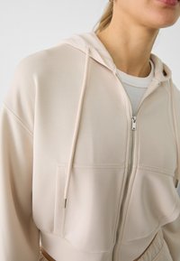 Sudadera de cremallera rosa claro, hecha de una tela suave, que presenta un bolsillo delantero, capucha con cordón y un diseño recortado con detalles de costura visibles.