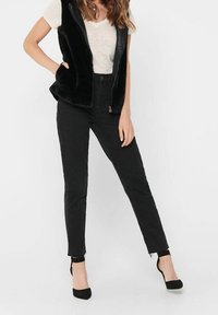 Svart fuskpäls väst över en ljusgrå skjorta, matchad med svarta skinny jeans och svarta klackskor med remmar. Släta texturer och åtsittande design.