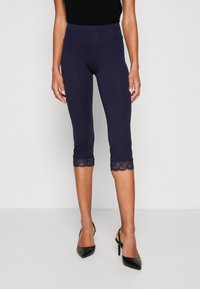 Leggings capri bleu marine avec finition en dentelle au bas, présentant une coupe ajustée et une texture lisse. Idéal pour des environnements décontractés ou relaxés.